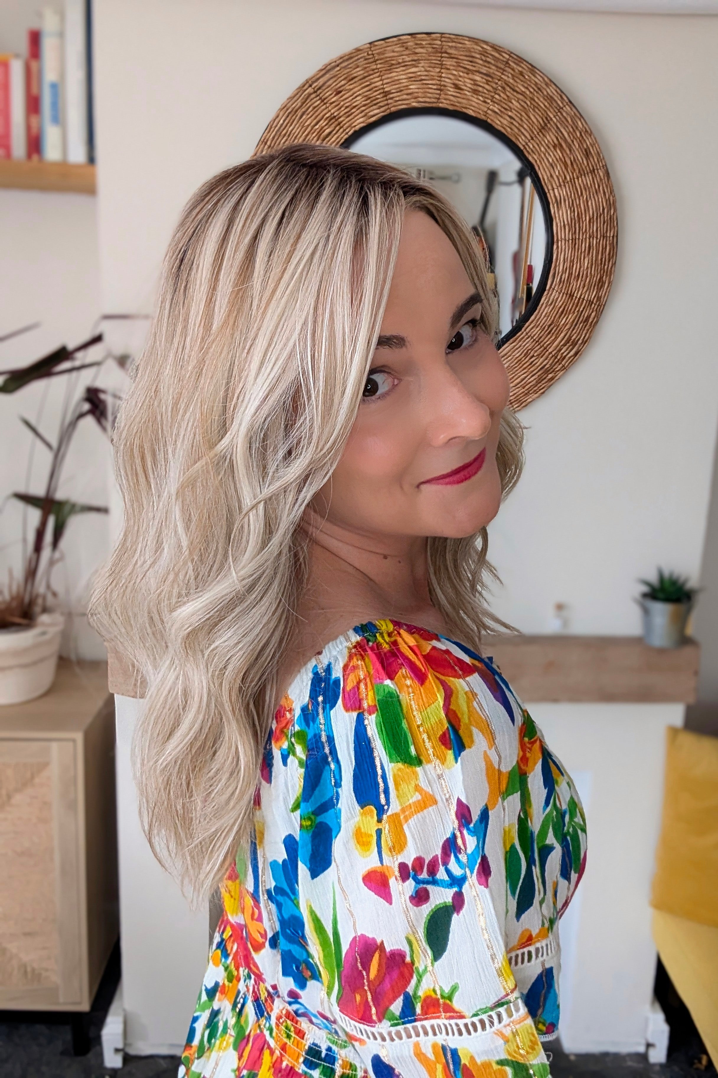 A woman with rooted blonde wavy hair wearing a vibrant, multicolored floral print top (Claudia by Belle Tress in Butterbeer R).