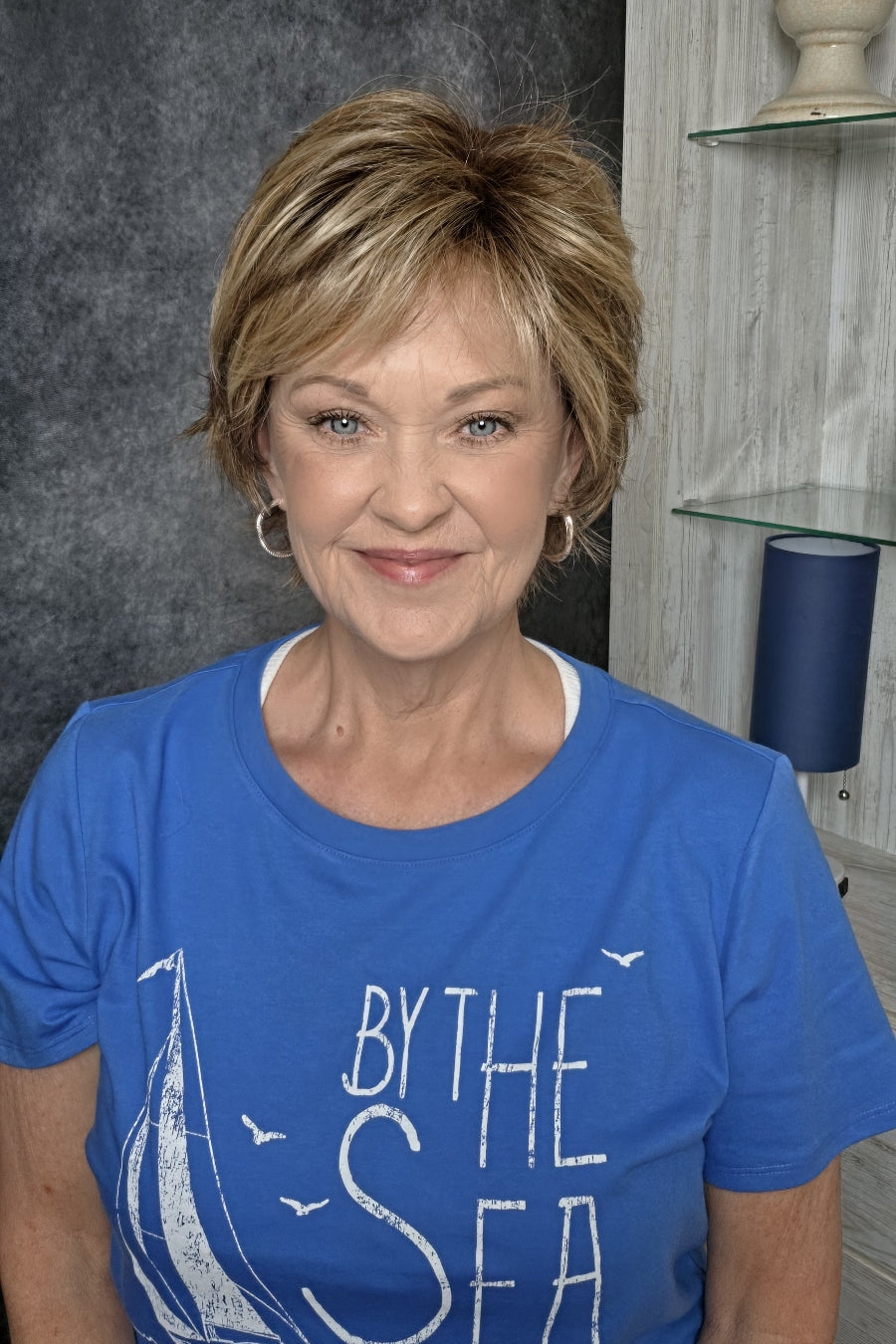 A smiling woman with short blonde wig wearing a blue graphic t-shirt, seated in a salon chair against a dark textured wall and light wood shelving. Avenue soft in Dark Sand R.