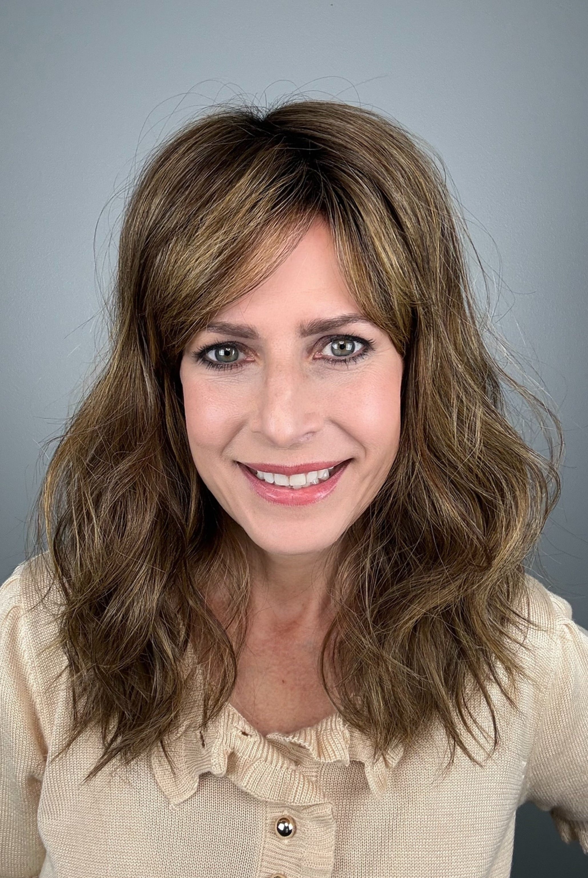 A woman with shoulder-length brown hair and bangs, wearing a light beige ruffled blouse (Great Vibe by Toni Brattin in color Brown Blonde).