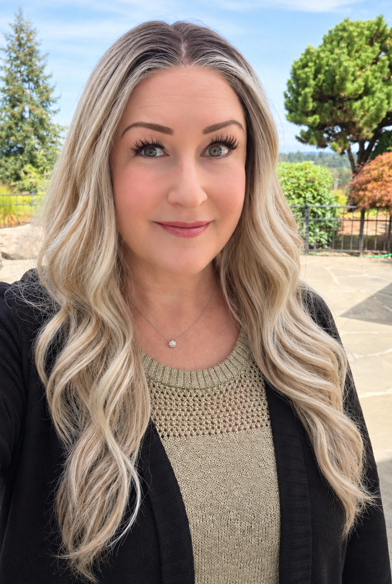 A woman wearing Bradley synthetic wig by PUSH in color Champagne Blonde Rooted is shown in a selfie-style outdoor portrait. She is wearing a black cardigan over a beige knit top, with a backyard pool, trees, and blue sky in the background.
