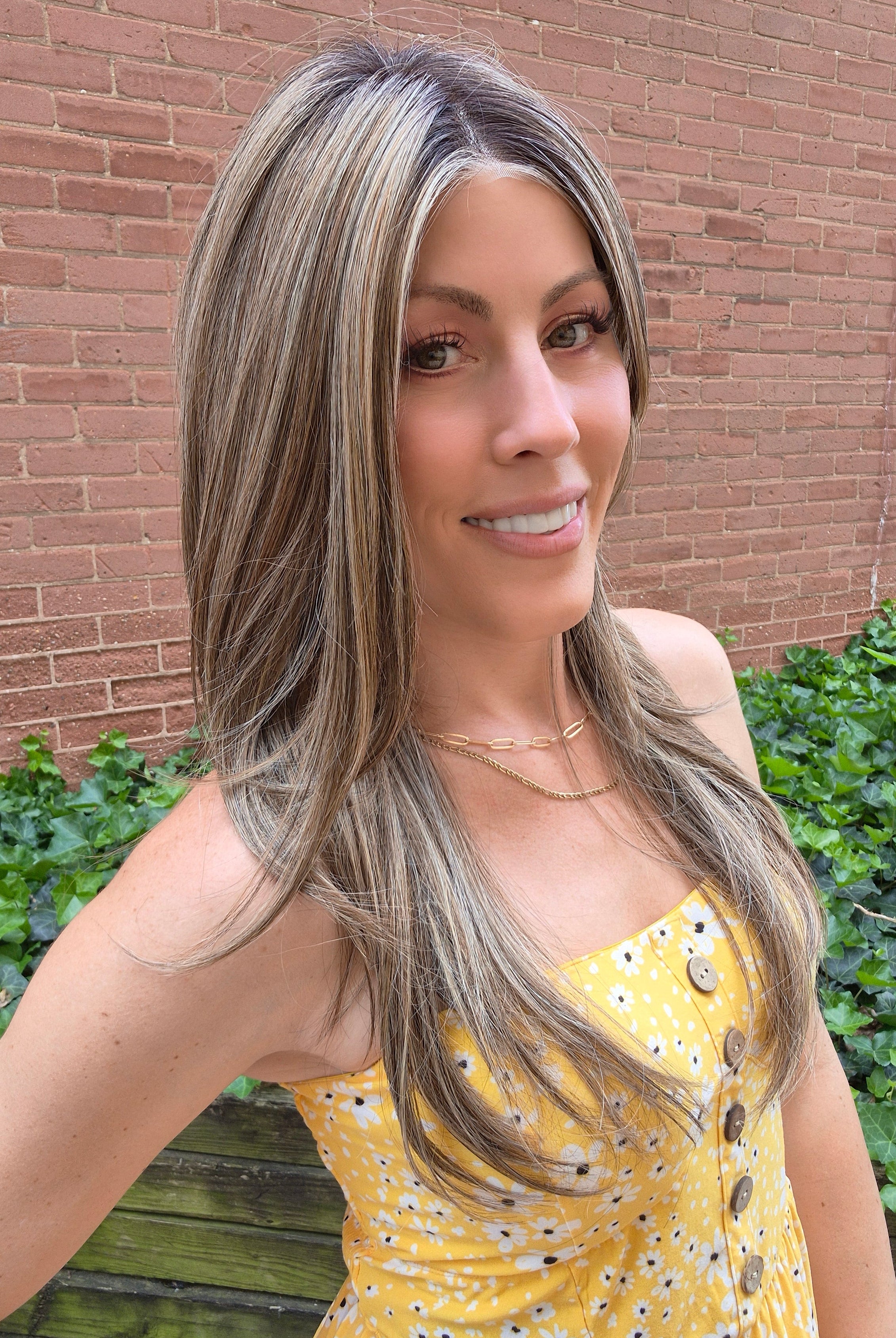 A woman with long highlighted brown and blonde wig wearing a yellow sleeveless top with black polka dots and brown buttons, photographed outdoors against a brick wall and green ivy. Scene Stealer by Raquel Welch in Shaded Cappuccino.
