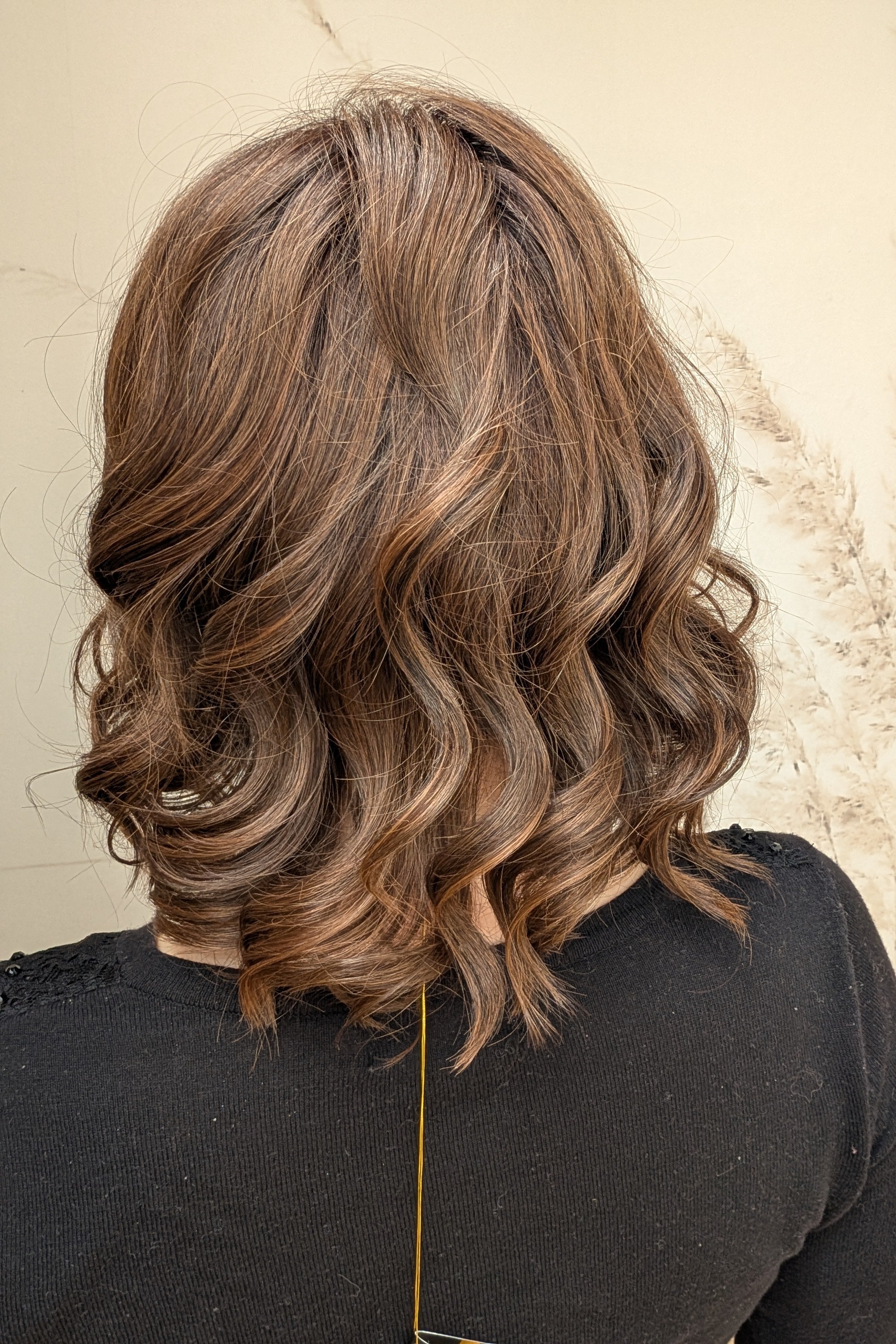 A woman with shoulder-length brown curly hair wearing a black long-sleeve top (Streep by Belle Tress in color Soft Amber Brown).