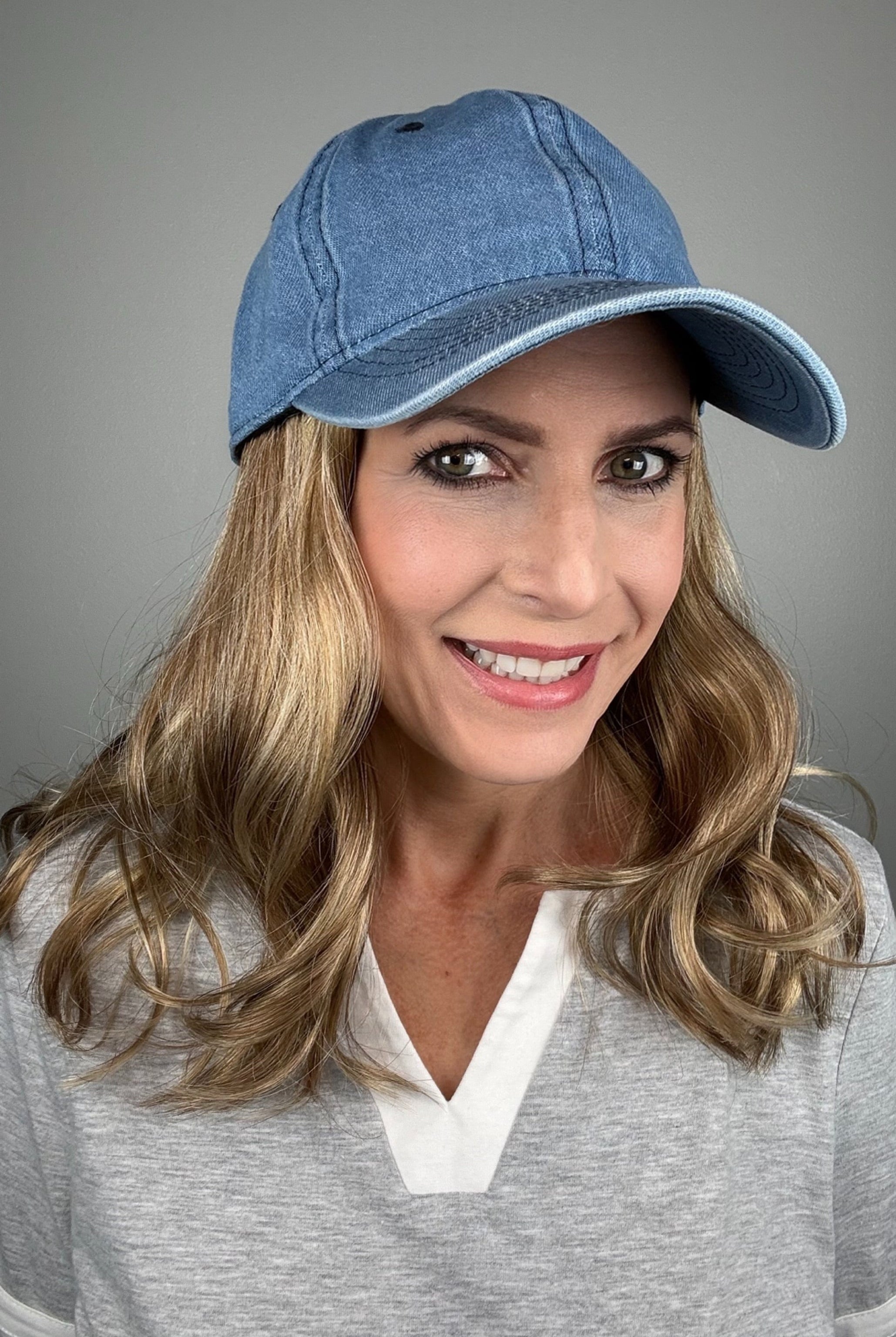 A woman with dark blonde hair wearing a blue denim baseball cap and a gray V-neck top (wearing Wavy Hat Denim in color 12/26H).