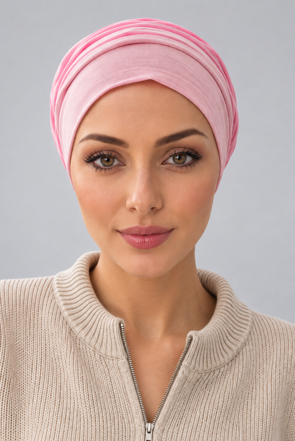 A woman wearing a rose melange turban-style cap, paired with a beige ribbed zip-up sweater, against a solid gray studio background. Front view