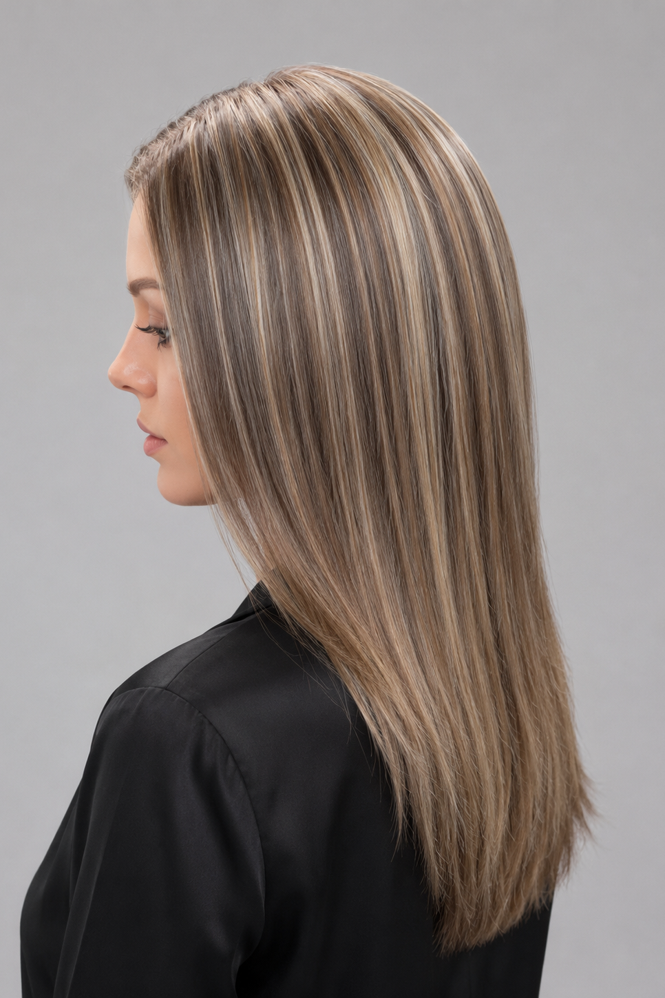 Woman wearing Evangeline heat friendly synthetic long wig in color cashmere bronde and a button down black blouse against a light grey background. Left side view.
