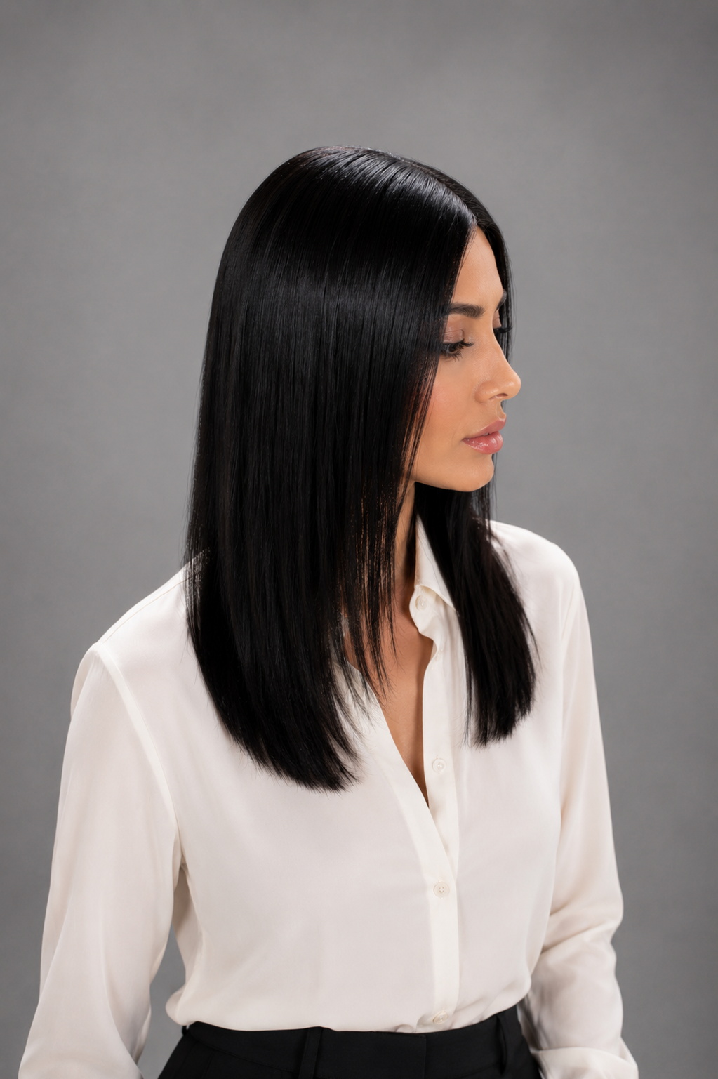 Woman wearing Jacey human hair wig by PUSH in color Noir with cream button down blouse and black pants with a light grey background. Right side view.