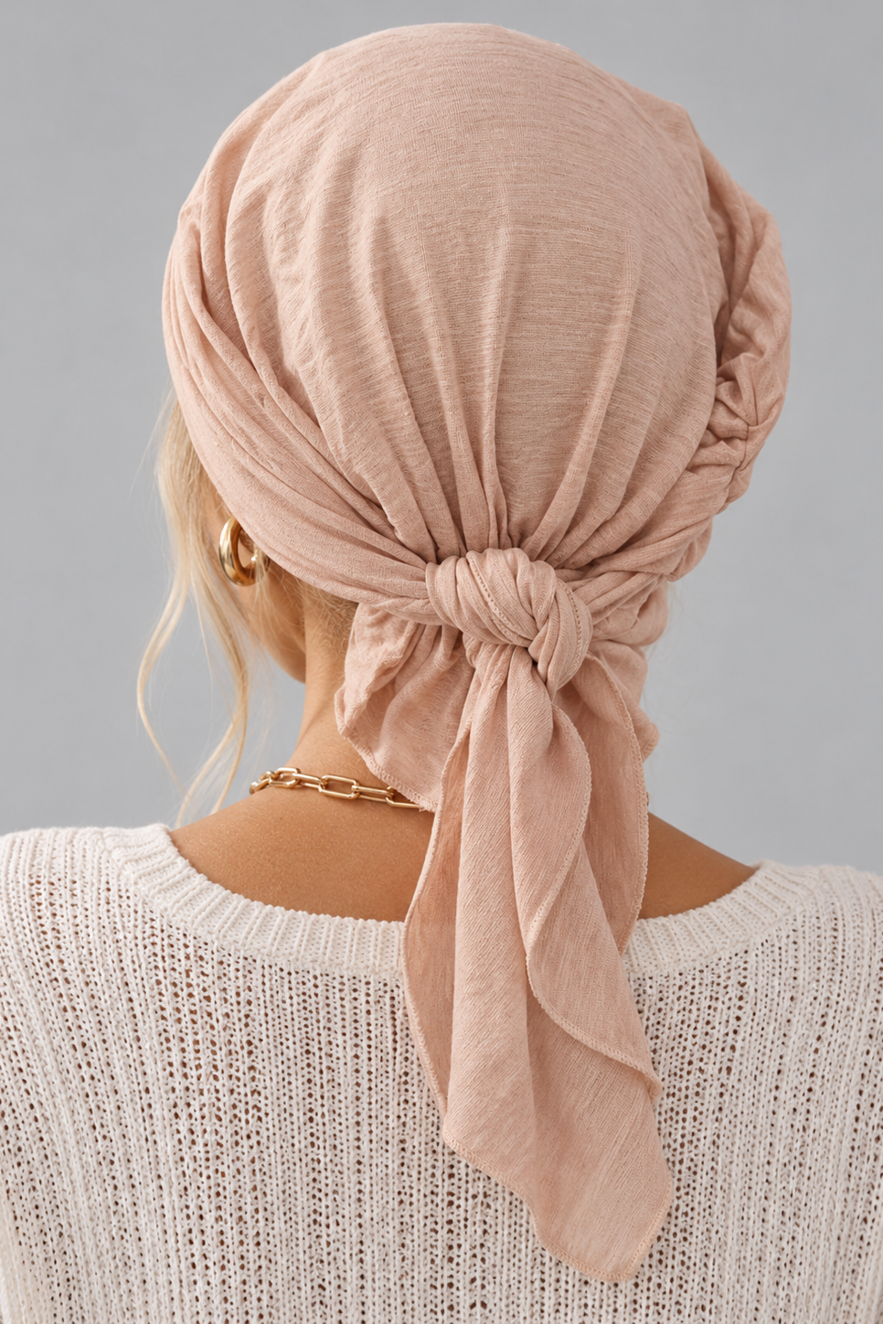 A woman wearing a soft amber melange bamboo head covering or turban-style cap, paired with a cream knit V-neck sweater and a gold chain necklace, against a plain light gray studio background. back view