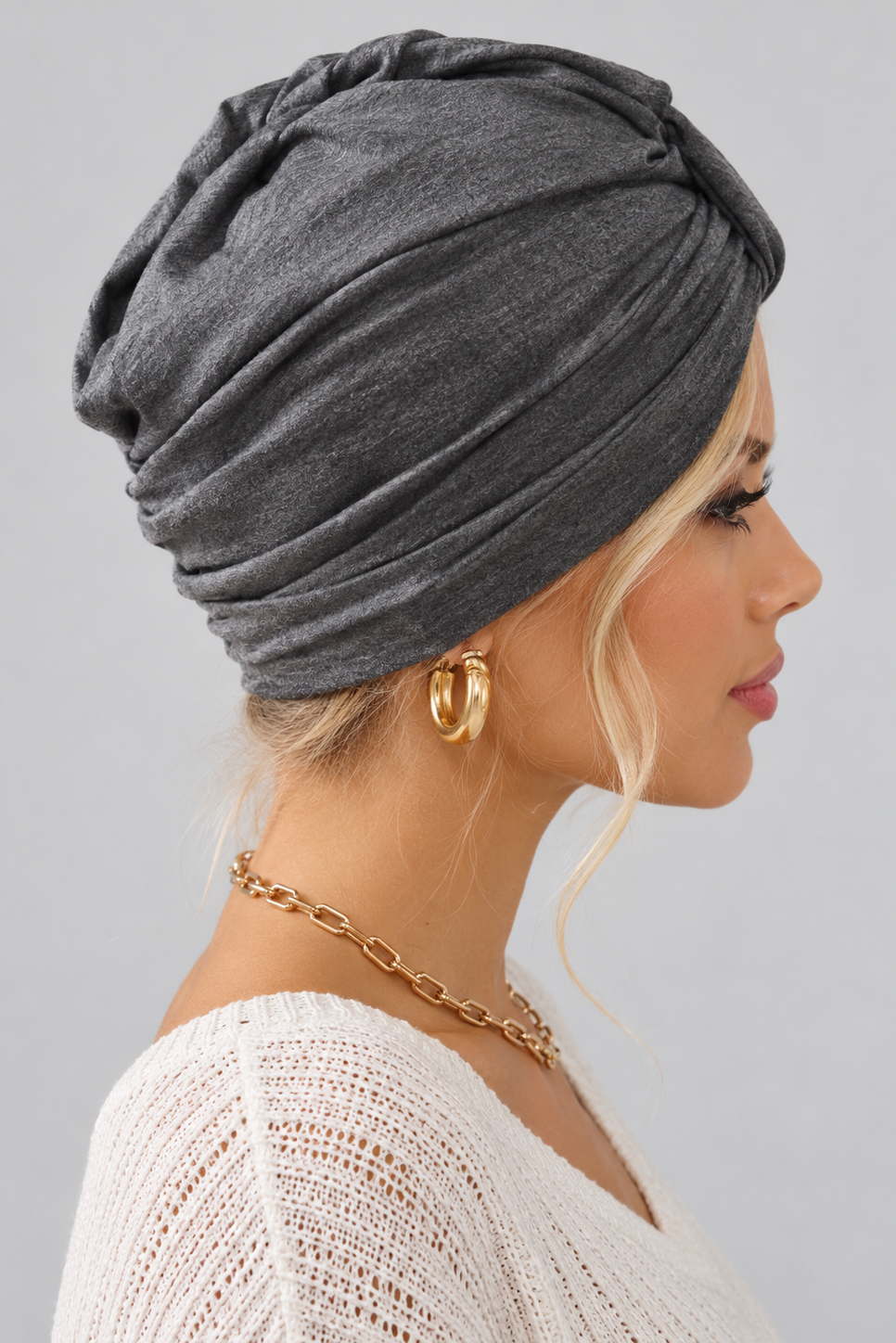 A woman wearing the Wisdom Bamboo Turban in color Anthracite Melange styled with gold hoop earrings and a gold chain necklace against a solid gray background. Right side view.