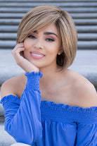 A woman with a short layered blonde bob wig named Abbey by Envy posing outdoors against gray concrete steps, wearing a bright blue off-the-shoulder top.