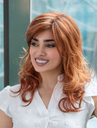 Woman wearing Alana synthetic wig by Envy in color Lighter Red wearing a white blouse in an outdoor setting.