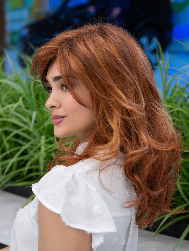 Woman wearing Alana synthetic wig by Envy in color Lighter Red wearing a white blouse in an outdoor setting. Left side.