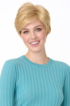 Blonde synthetic pixie wig with long bangs on a smiling woman in a blue ribbed shirt.
