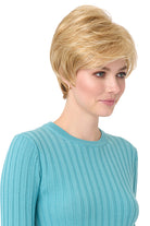 Blonde synthetic Angel by Envy pixie wig with long bangs on a woman in a blue top.