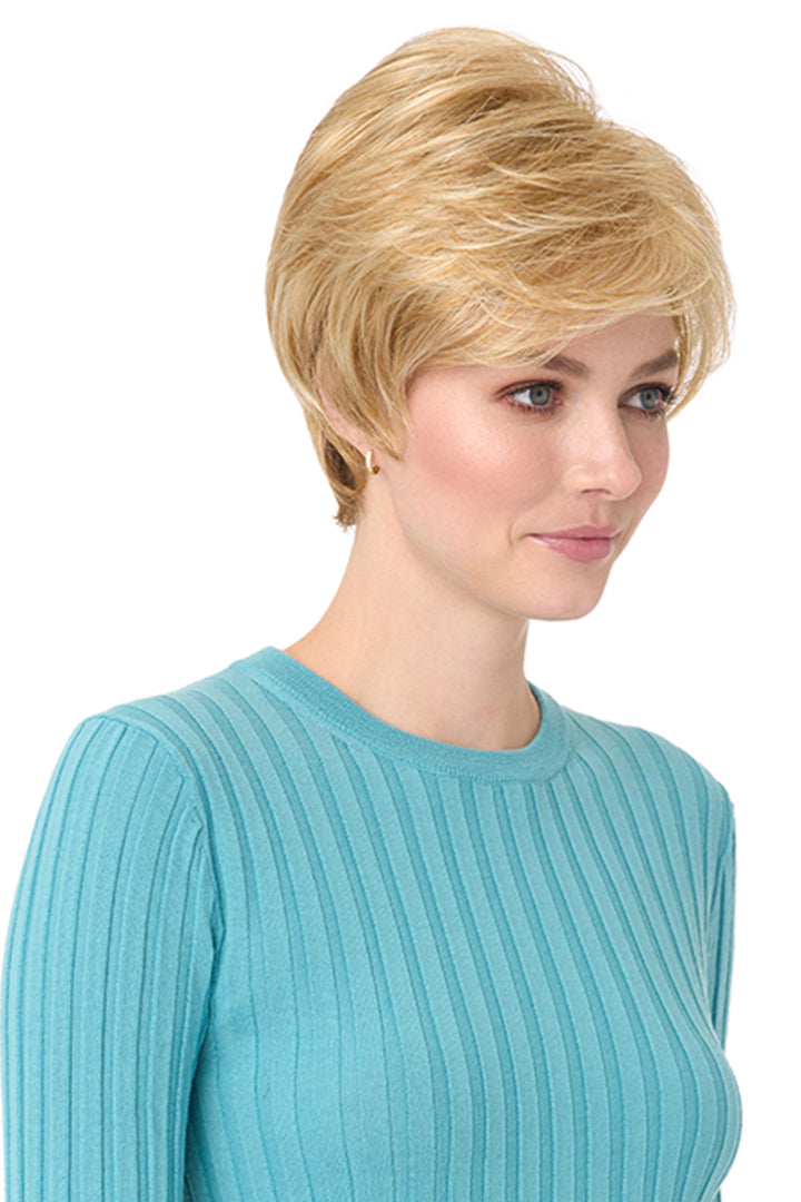 Blonde synthetic Angel by Envy pixie wig with long bangs on a woman in a blue top.