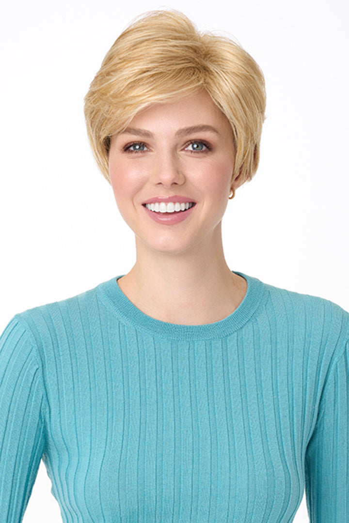 Blonde synthetic pixie wig with long bangs on a smiling woman in a blue ribbed shirt.