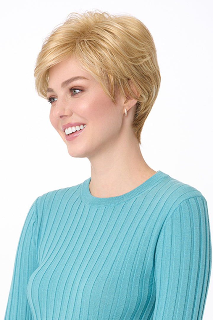 Blonde synthetic pixie wig with long bangs on a smiling woman in a blue ribbed top.