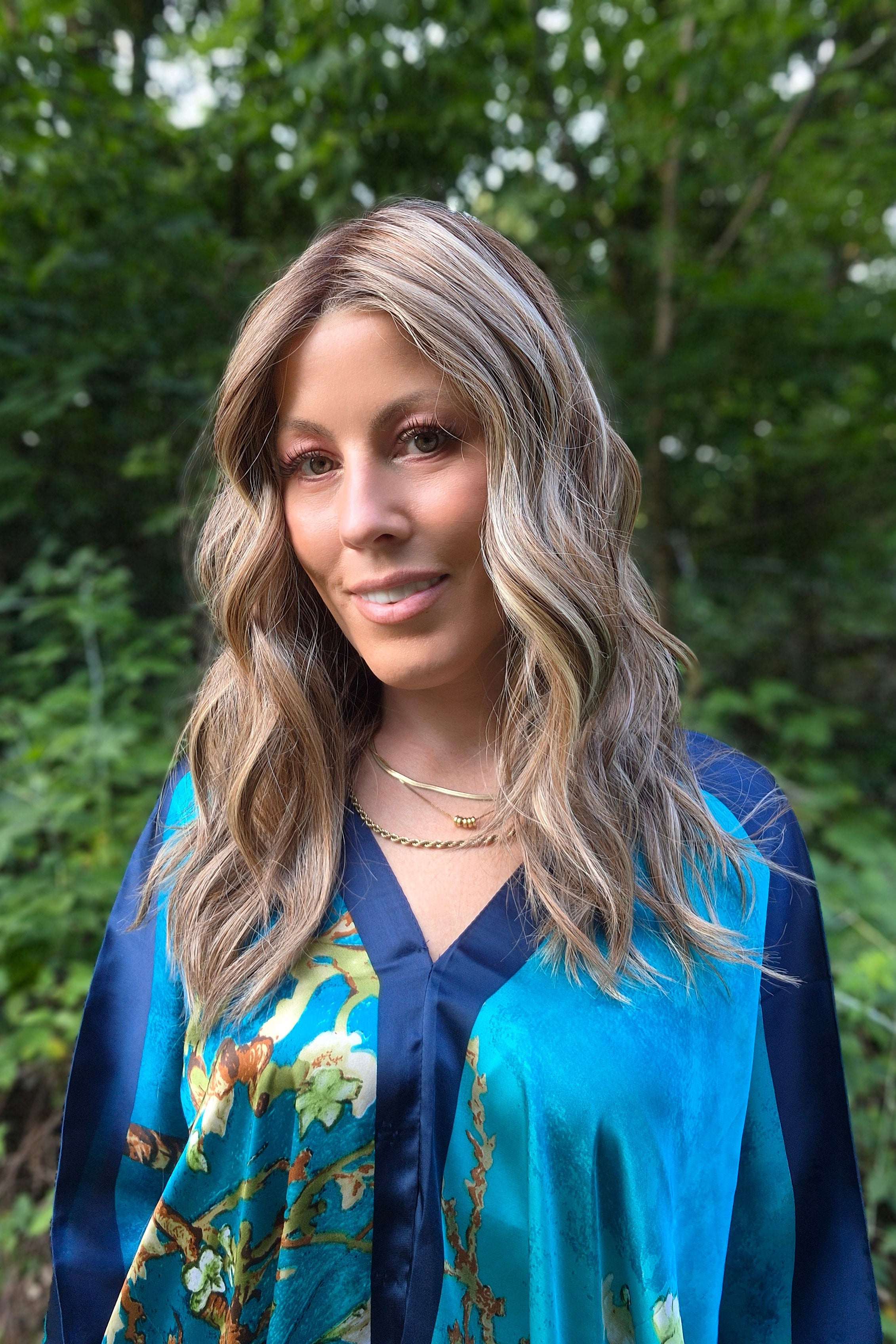 A woman with blonde-highlighted hair wearing a blue and teal floral kimono-style top (wearing Bella by Belle Tress in Brown Sugar Cookie R).
