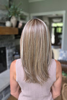 A smiling woman with shoulder-length layered blonde hair and bangs, wearing a sleeveless taupe ribbed top (Bailey by Belle Tress in Brown Sugar Cookie R).