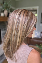 A smiling woman with shoulder-length layered blonde hair and bangs, wearing a sleeveless taupe ribbed top (Bailey by Belle Tress in Brown Sugar Cookie R).