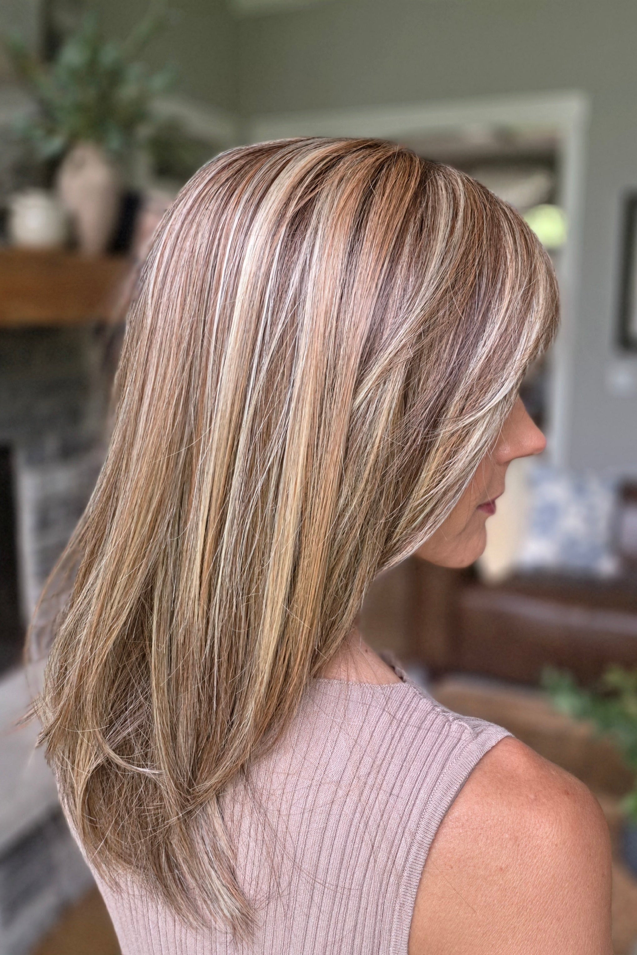 A smiling woman with shoulder-length layered blonde hair and bangs, wearing a sleeveless taupe ribbed top (Bailey by Belle Tress in Brown Sugar Cookie R).