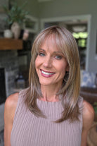 A smiling woman with shoulder-length layered blonde hair and bangs, wearing a sleeveless taupe ribbed top (Bailey by Belle Tress in Brown Sugar Cookie R).