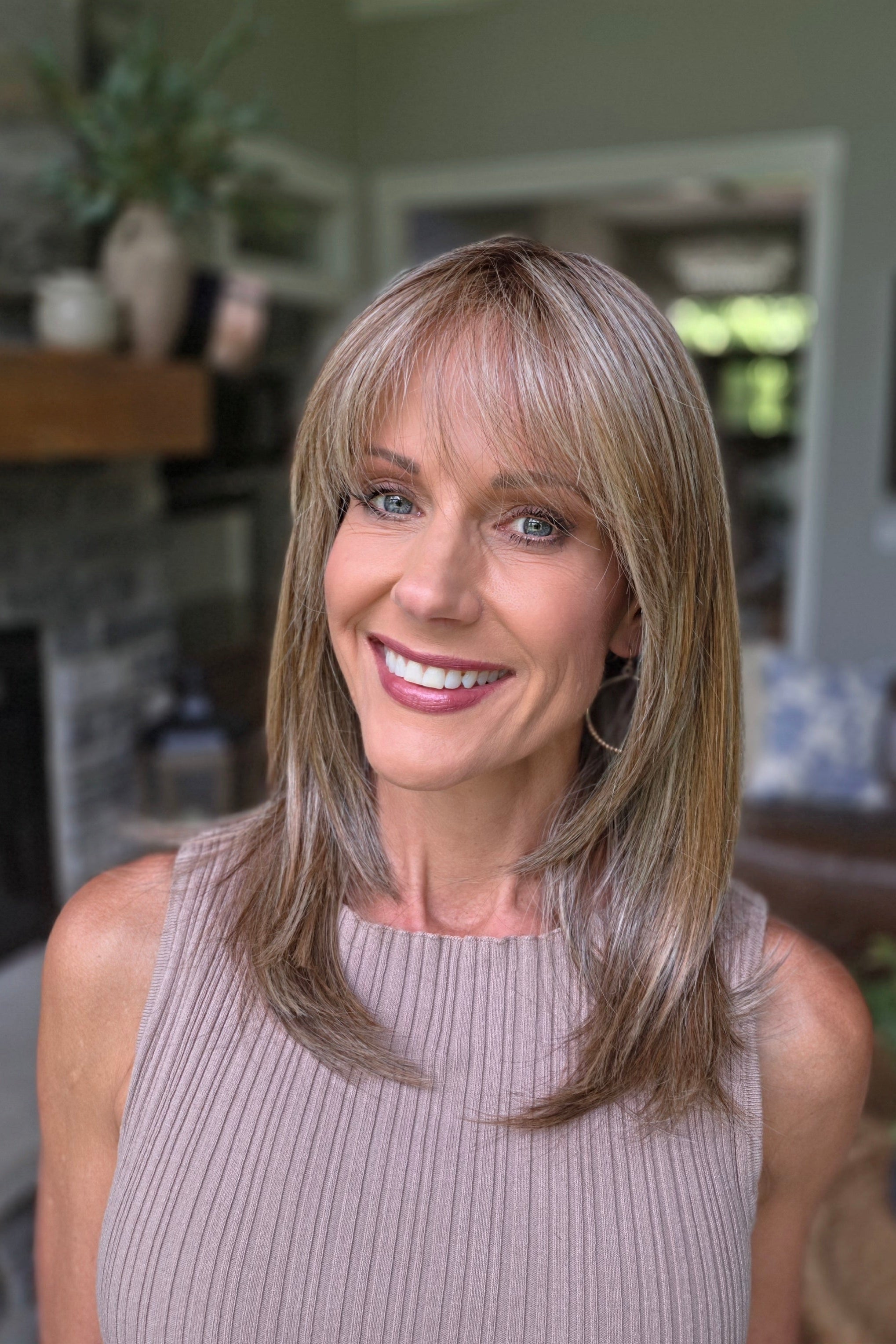 A smiling woman with shoulder-length layered blonde hair and bangs, wearing a sleeveless taupe ribbed top (Bailey by Belle Tress in Brown Sugar Cookie R).