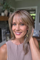 A smiling woman with shoulder-length layered blonde hair and bangs, wearing a sleeveless taupe ribbed top (Bailey by Belle Tress in Brown Sugar Cookie R).