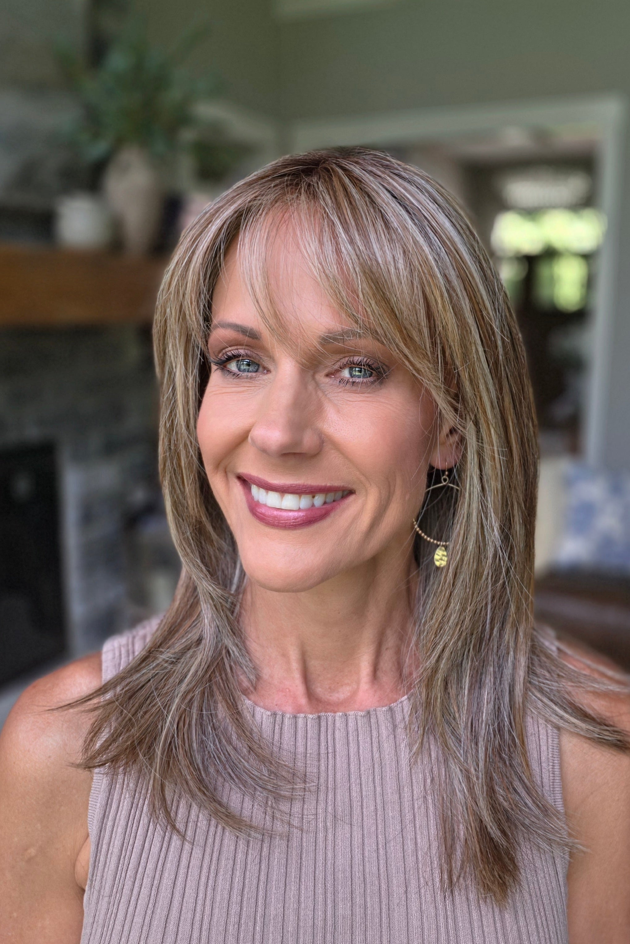 A smiling woman with shoulder-length layered blonde hair and bangs, wearing a sleeveless taupe ribbed top (Bailey by Belle Tress in Brown Sugar Cookie R).