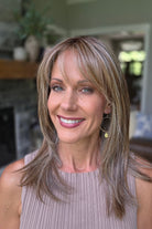 A smiling woman with shoulder-length layered blonde hair and bangs, wearing a sleeveless taupe ribbed top (Bailey by Belle Tress in Brown Sugar Cookie R).