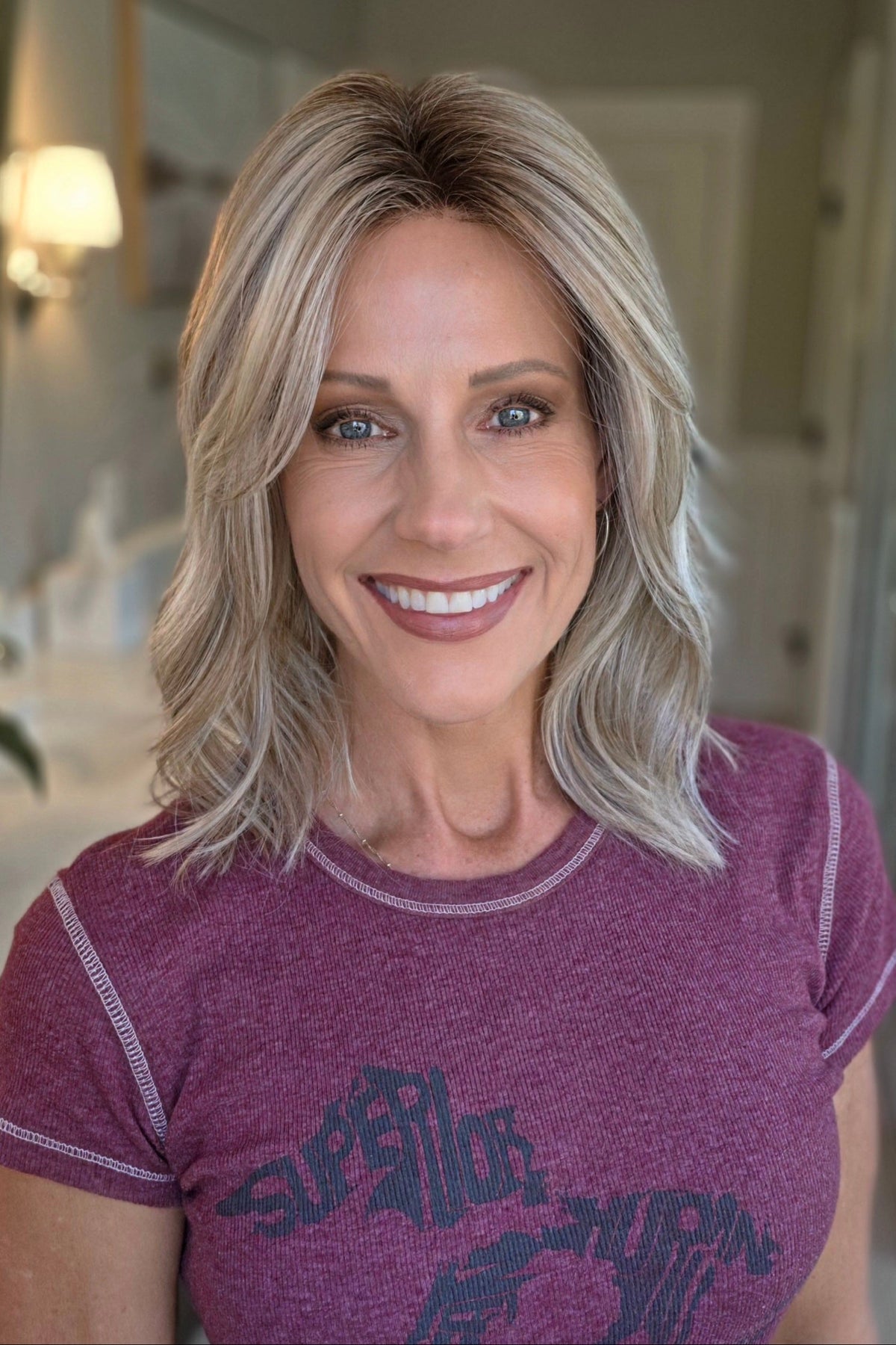 A smiling woman with shoulder-length wavy blonde wig (Monaco by Belle Tress in color Milkshake Blonde R).