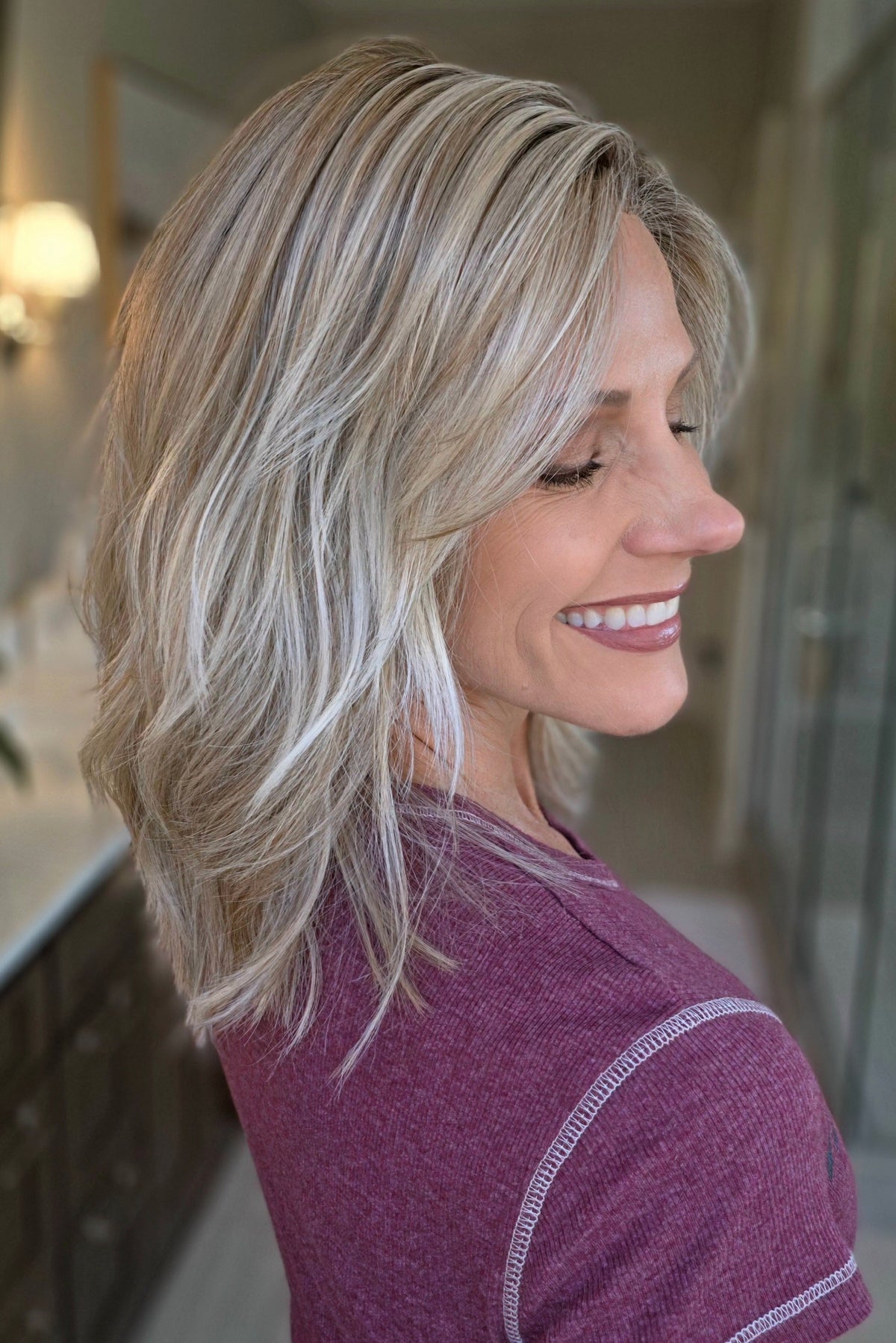 A smiling woman with shoulder-length wavy blonde wig (Monaco by Belle Tress in color Milkshake Blonde R).