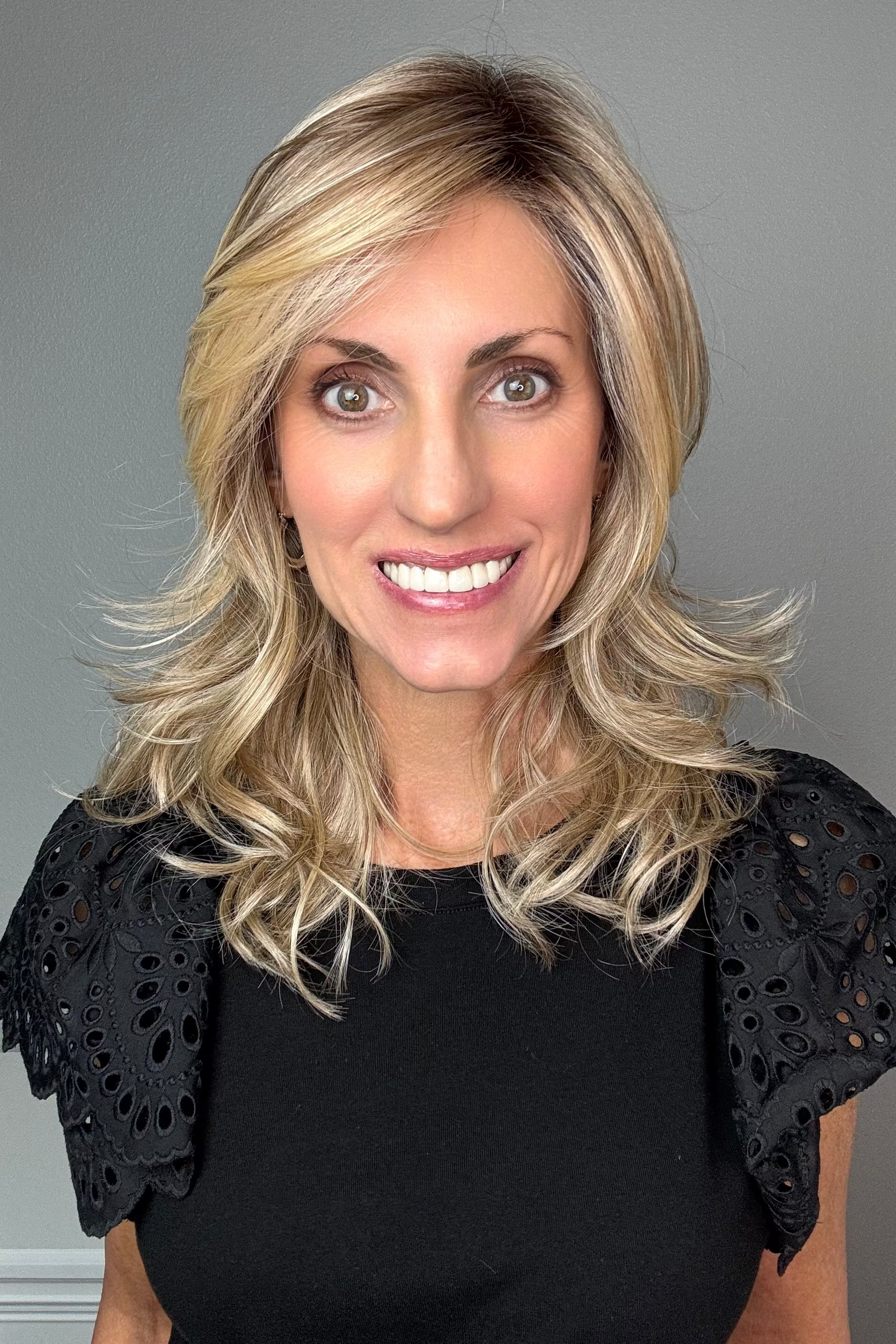 A smiling woman with shoulder-length layered blonde wig (Munich by Belle Tress in Raw Sugar Blonde).