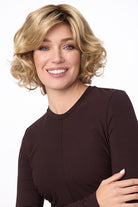 Smiling woman wearing a mid-length, curly blonde synthetic wig with side-swept bangs.