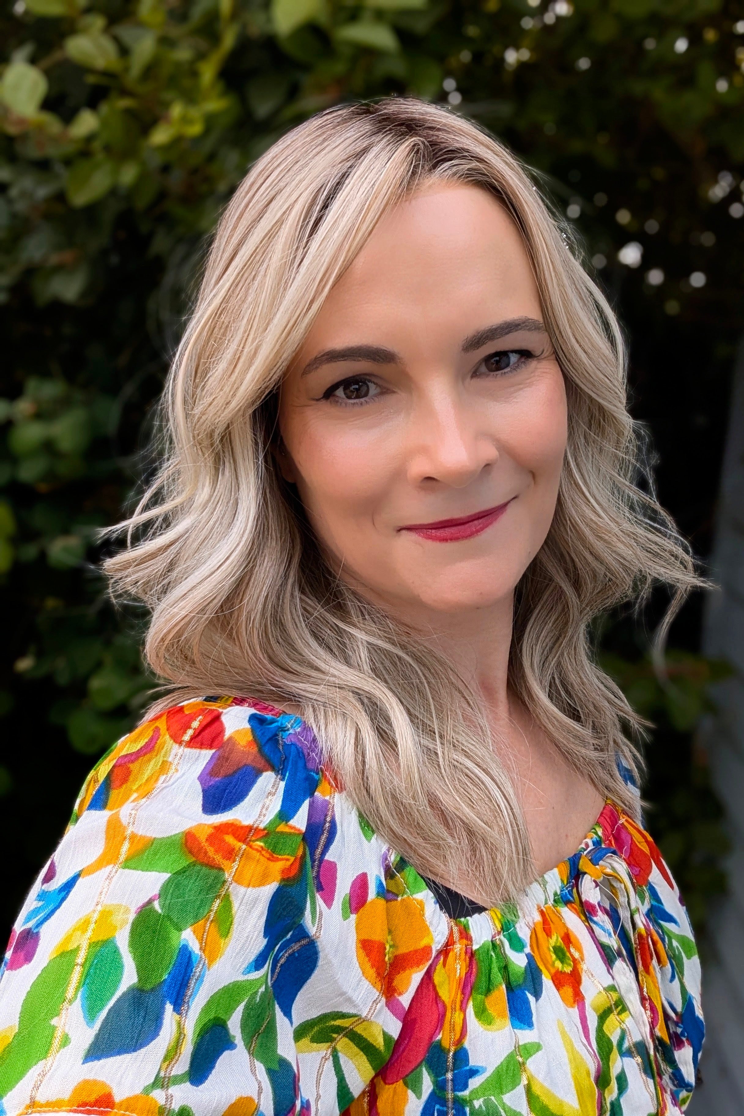 A woman with rooted blonde wavy hair wearing a vibrant, multicolored floral print top (Claudia by Belle Tress in Butterbeer R).