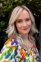 A woman with rooted blonde wavy hair wearing a vibrant, multicolored floral print top (Claudia by Belle Tress in Butterbeer R).