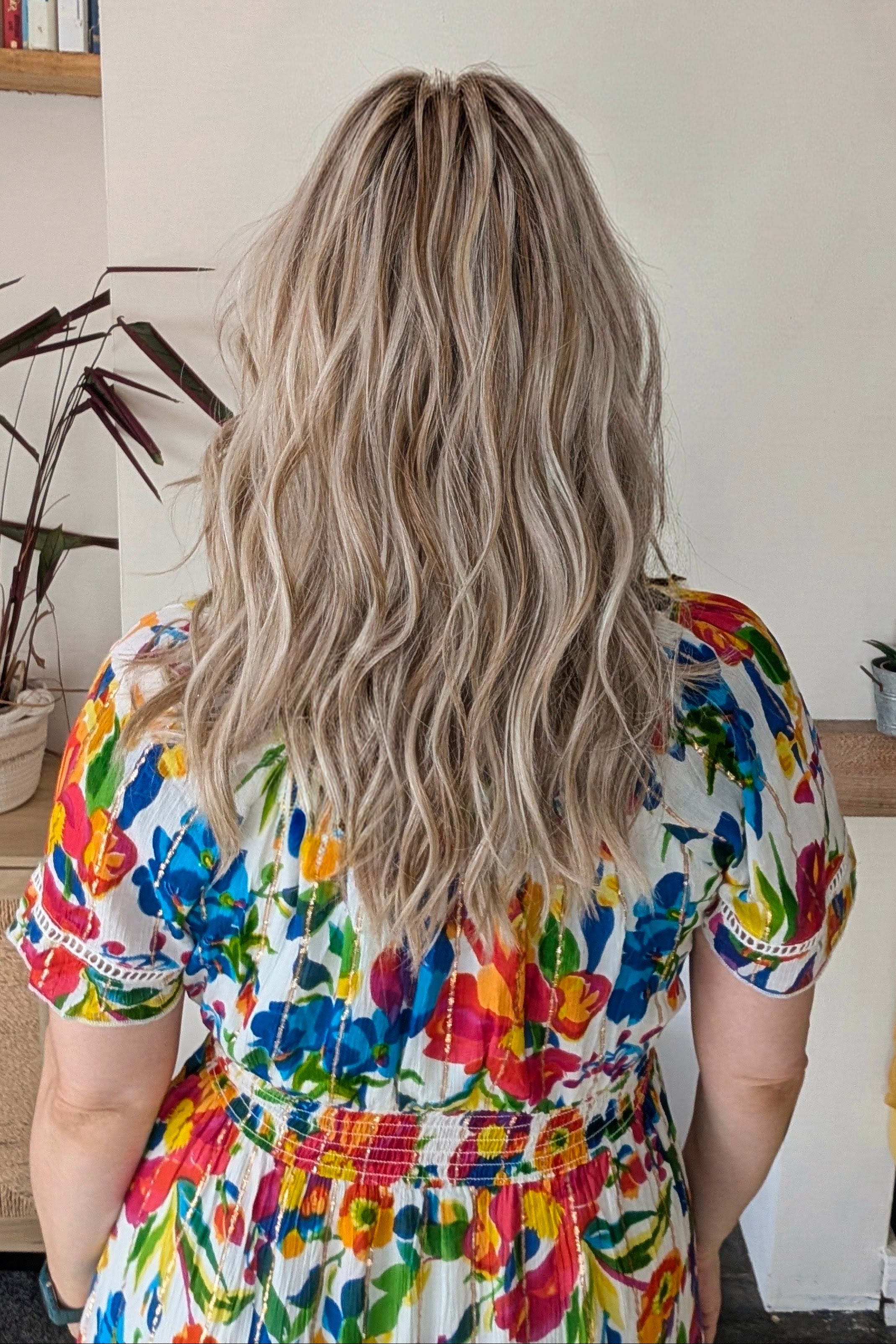A woman with rooted blonde wavy hair wearing a vibrant, multicolored floral print top (Claudia by Belle Tress in Butterbeer R).