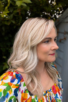 A woman with rooted blonde wavy hair wearing a vibrant, multicolored floral print top (Claudia by Belle Tress in Butterbeer R).