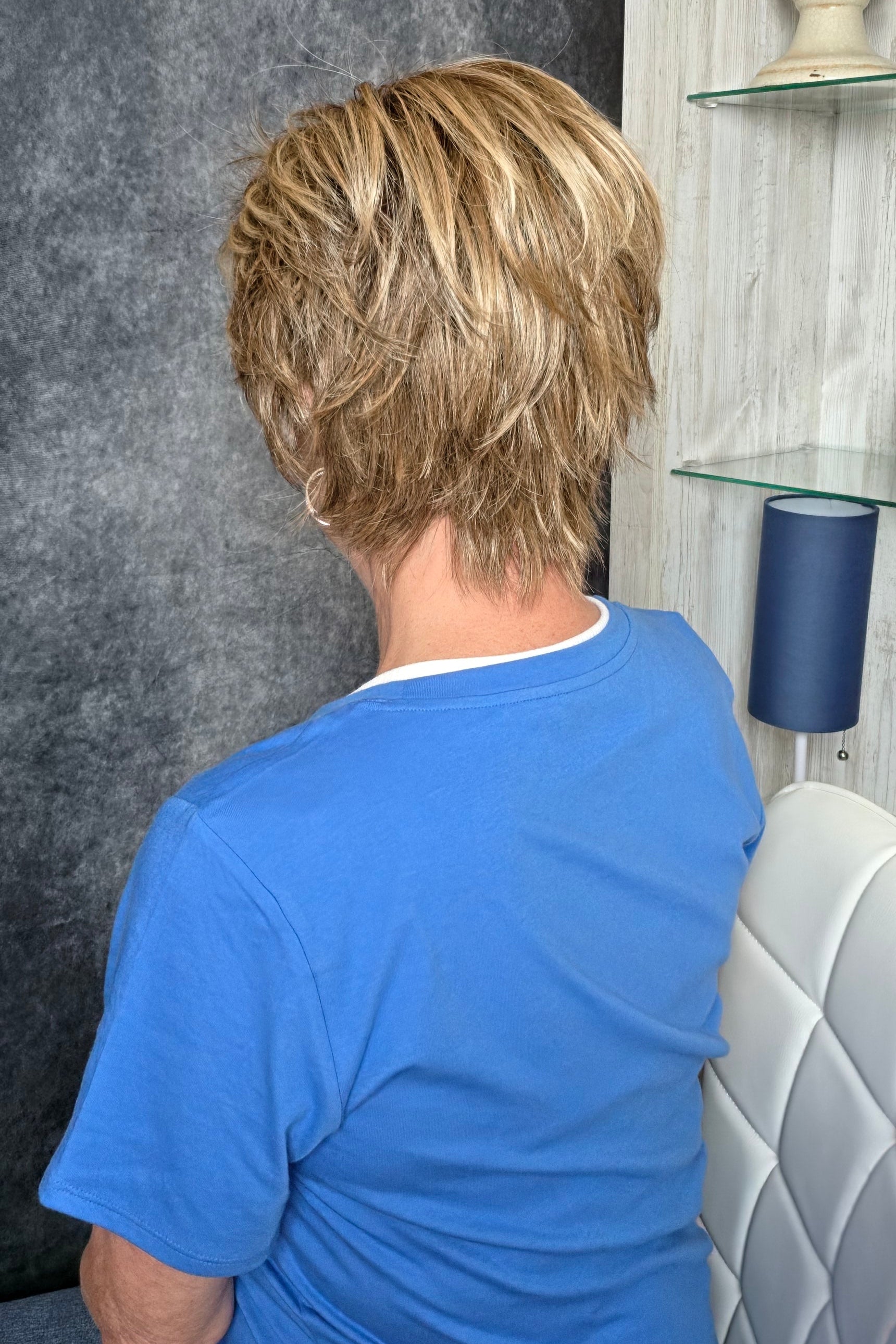 A smiling woman with short blonde wig wearing a blue graphic t-shirt, seated in a salon chair against a dark textured wall and light wood shelving. Avenue soft in Dark Sand R.