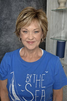 A smiling woman with short blonde wig wearing a blue graphic t-shirt, seated in a salon chair against a dark textured wall and light wood shelving. Avenue soft in Dark Sand R.