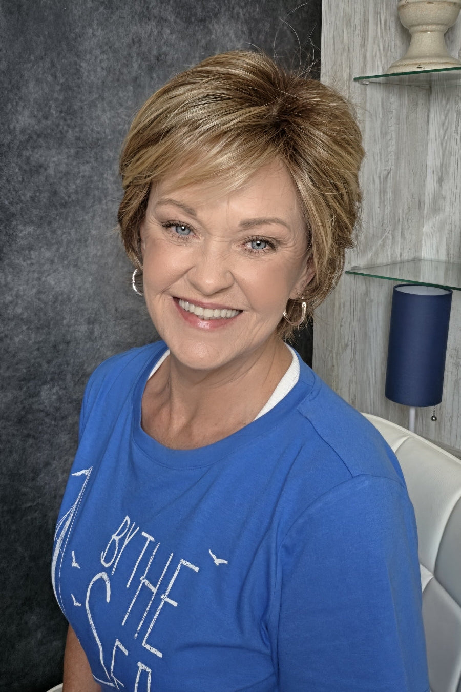 A smiling woman with short blonde wig wearing a blue graphic t-shirt, seated in a salon chair against a dark textured wall and light wood shelving. Avenue soft in Dark Sand R.