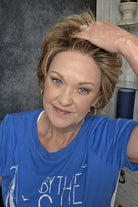 A smiling woman with short blonde wig wearing a blue graphic t-shirt, seated in a salon chair against a dark textured wall and light wood shelving. Avenue soft in Dark Sand R.