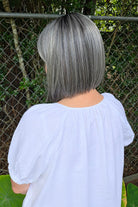A woman with shoulder-length gray wig wearing a white short-sleeve blouse with puff sleeves, standing outdoors in front of a chain-link fence and green foliage. Miss Comfort by Ellen Wille in Salt and Pepper Frosted.