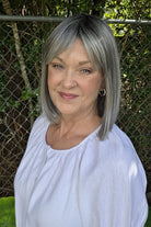 A woman with shoulder-length gray wig wearing a white short-sleeve blouse with puff sleeves, standing outdoors in front of a chain-link fence and green foliage. Miss Comfort by Ellen Wille in Salt and Pepper Frosted.