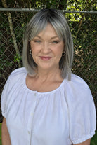 A woman with shoulder-length gray wig wearing a white short-sleeve blouse with puff sleeves, standing outdoors in front of a chain-link fence and green foliage. Miss Comfort by Ellen Wille in Salt and Pepper Frosted. 