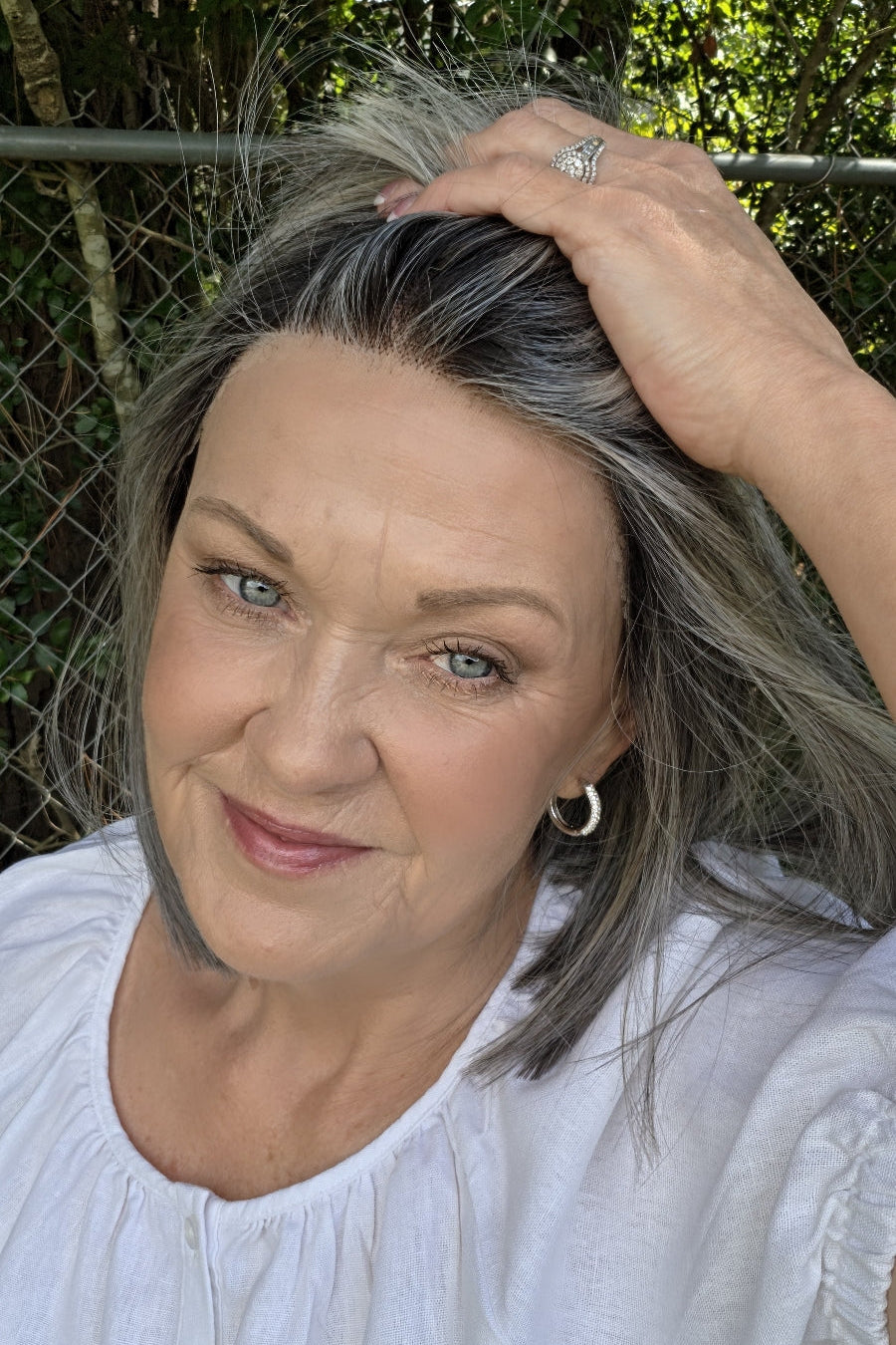 A woman with shoulder-length gray wig wearing a white short-sleeve blouse with puff sleeves, standing outdoors in front of a chain-link fence and green foliage. Miss Comfort by Ellen Wille in Salt and Pepper Frosted.