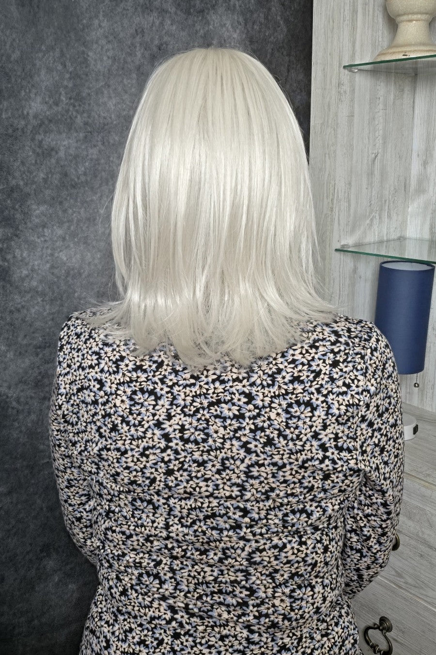 A woman with shoulder-length silver wig wearing a black and white floral long-sleeve top, photographed indoors against a dark textured wall and light wood shelving. Lisa by Envy in Light Gray.