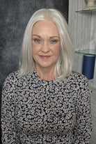 A woman with shoulder-length silver wig wearing a black and white floral long-sleeve top, photographed indoors against a dark textured wall and light wood shelving. Lisa by Envy in Light Gray