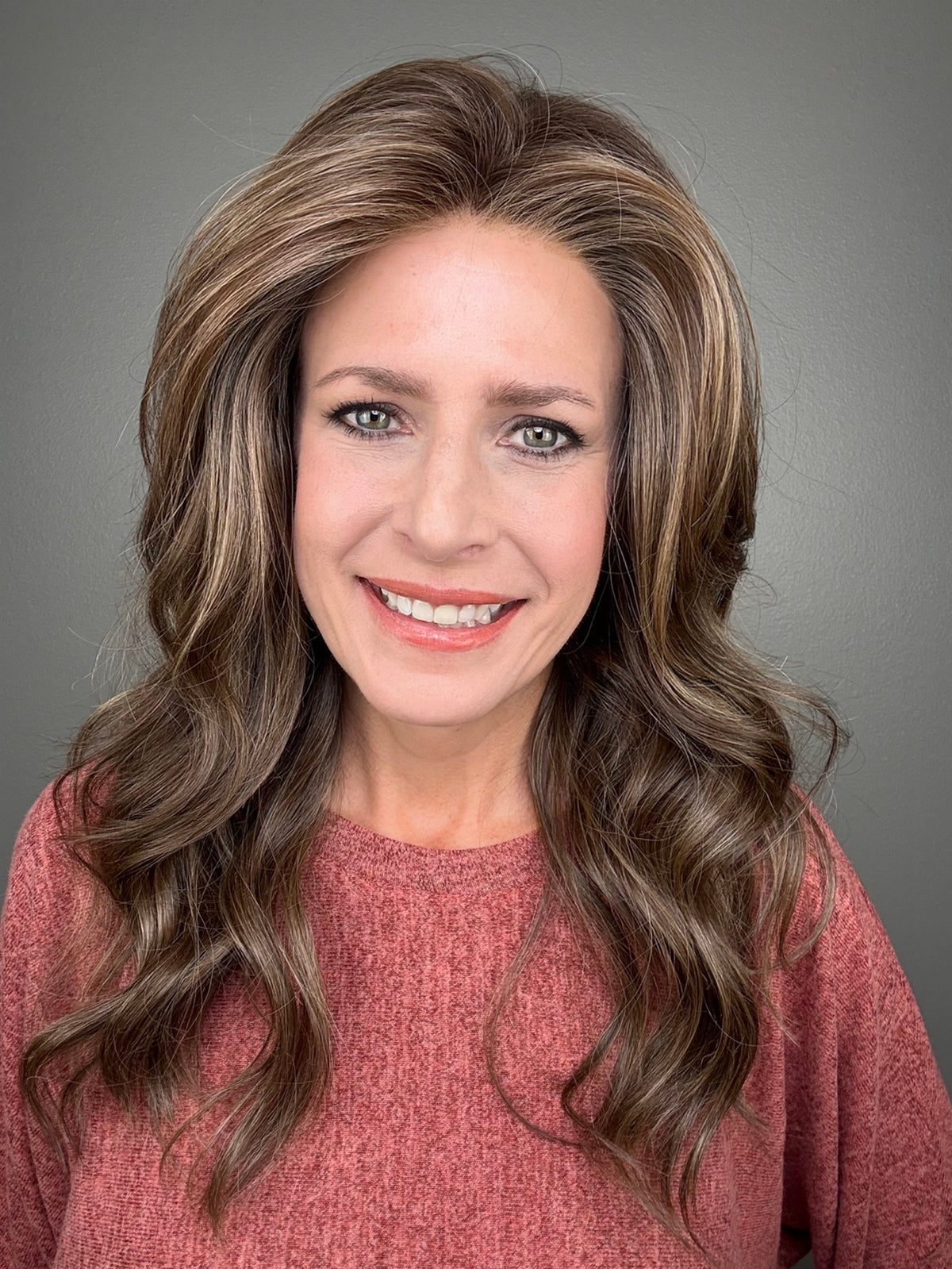 A smiling woman with long, wavy brown wig with blonde highlights (Fiona Lite by Jon Renau in color Mousse Cake).