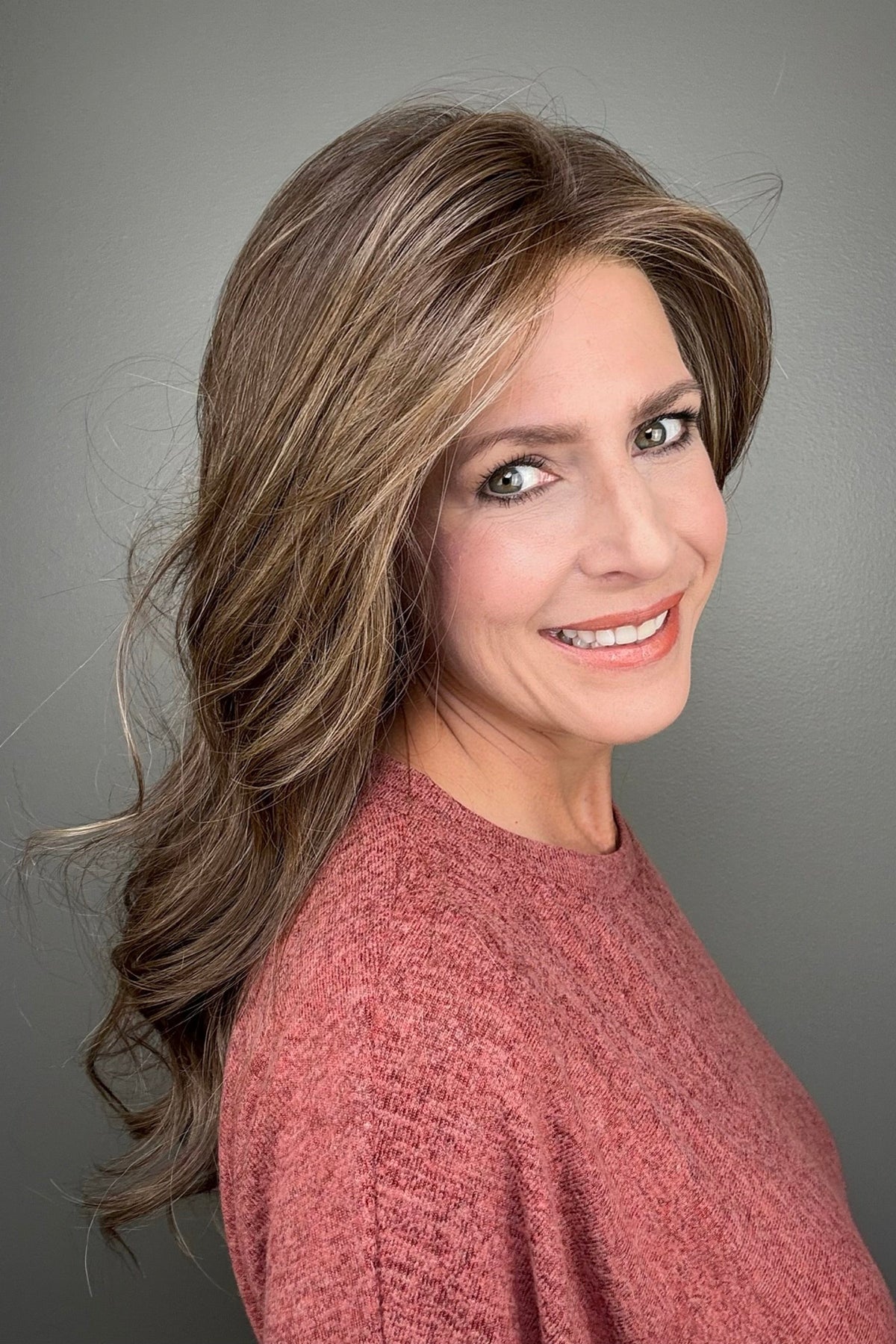 A smiling woman with long, wavy brown wig with blonde highlights (Fiona Lite by Jon Renau in color Mousse Cake).