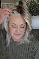 A smiling woman with shoulder-length layered blonde hair wearing an olive green cable-knit sweater (Sloane by Jon Renau in Catalina Blonde).